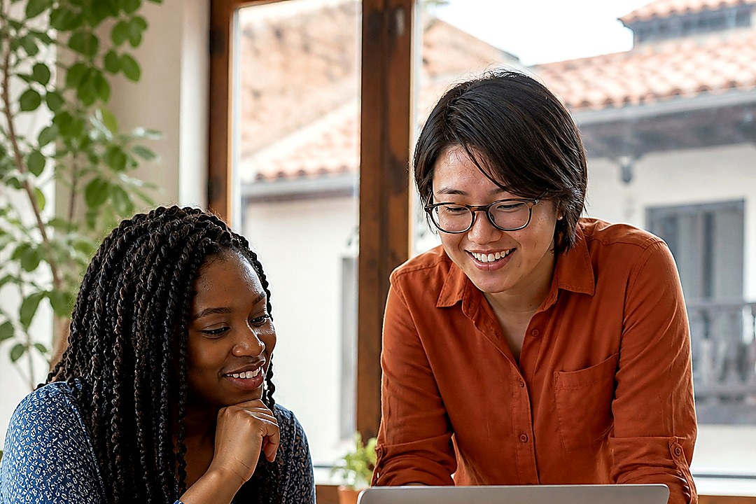 Two adult women collaborate around a laptop in a bright modern workspace.